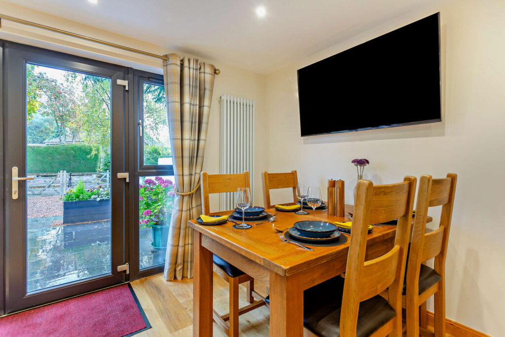 Dining area at Highfield Lodge with wooden table for four, wall-mounted TV and glazed doors opening to the patio.