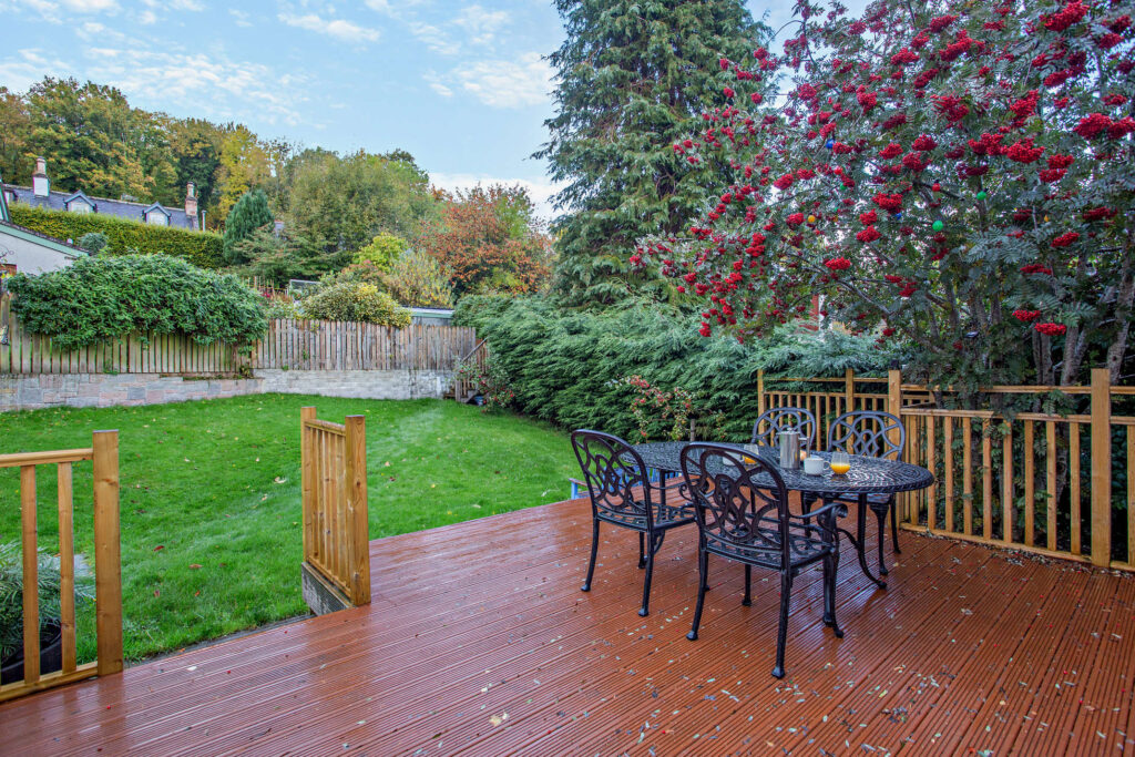 Private deck and lawned garden at Highfield Lodge with outdoor dining table beneath mature trees.
