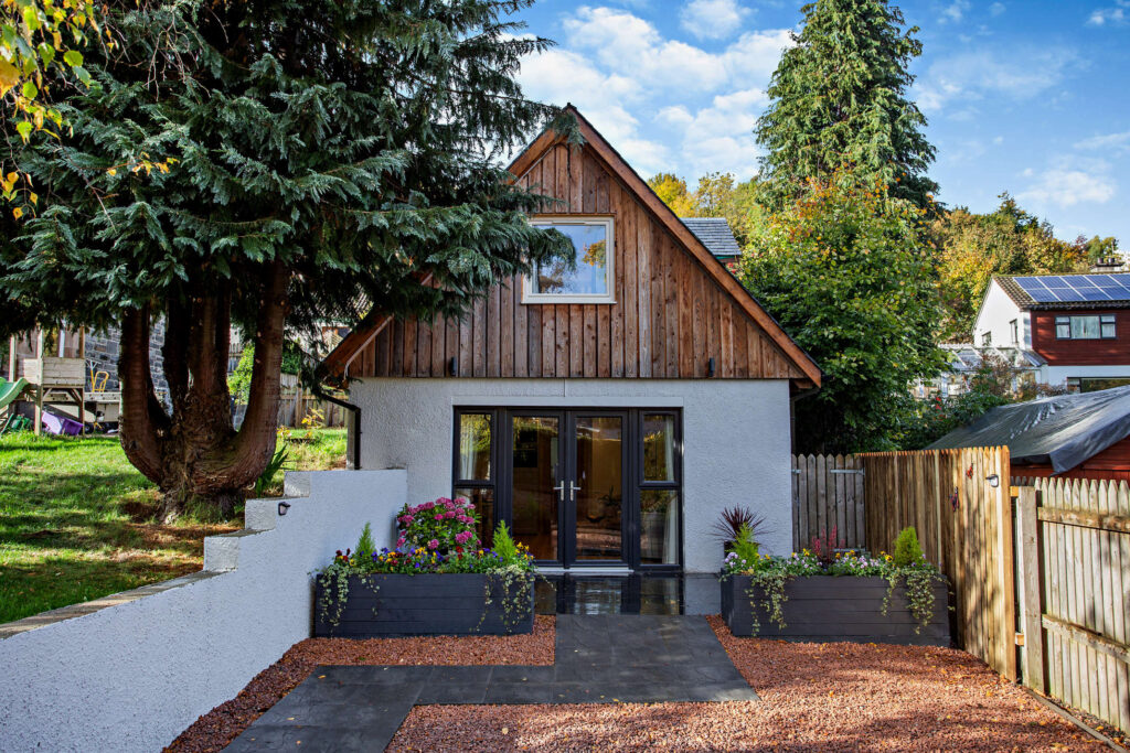 Front exterior of Highfield Lodge with timber-clad upper floor, patio doors, patio area and gravel driveway.