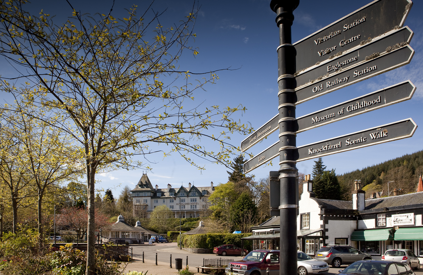 Directional signpost in Strathpeffer Square, with the Highland Hotel and village buildings in the background.