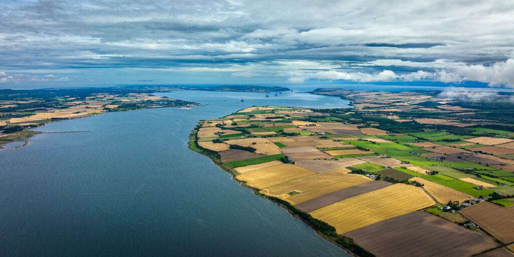 Aerial view east along the Cromarty Firth towards the Port of Nigg and the Sutors, with patchwork farmland along the shoreline and oil rigs in the distance.